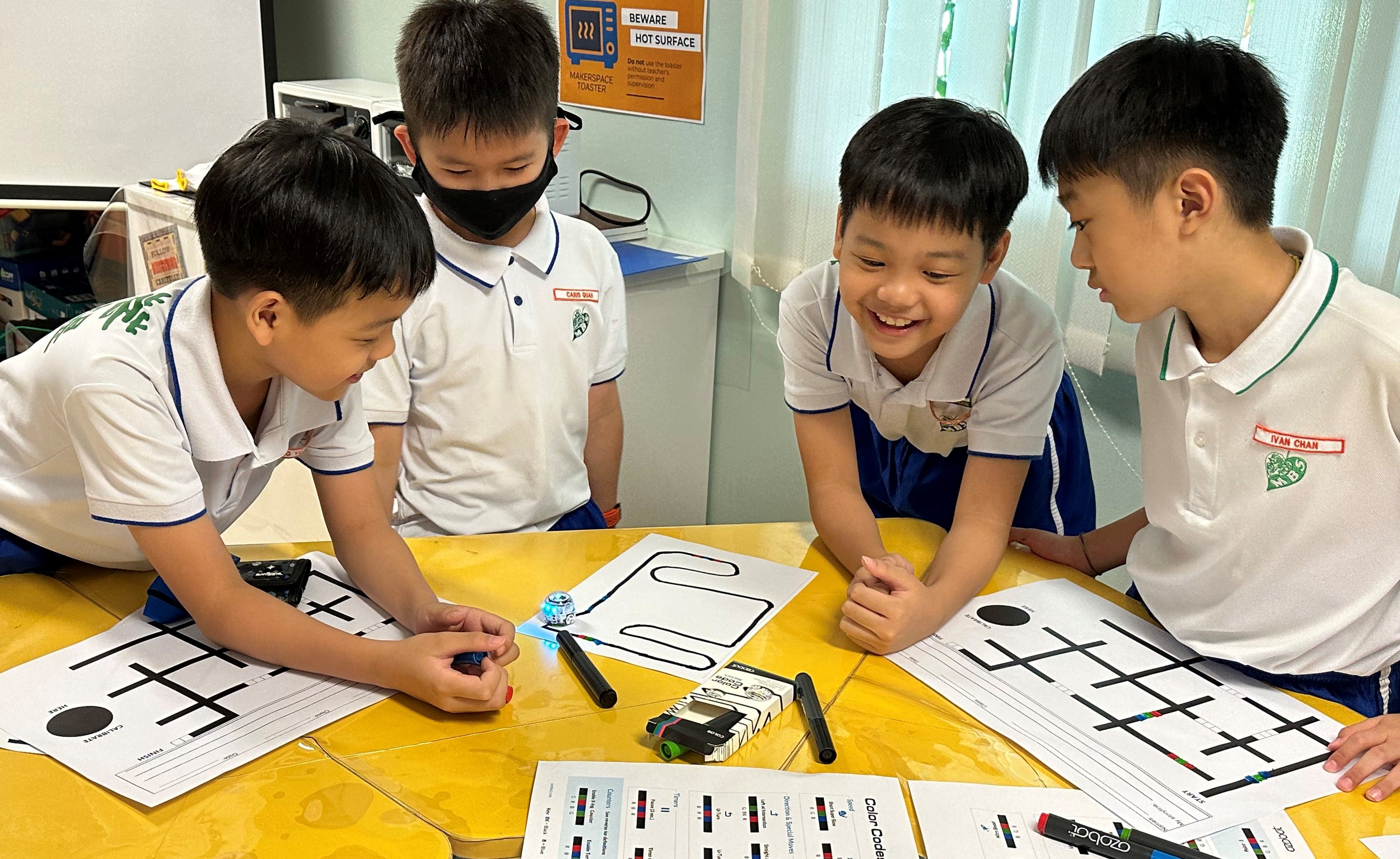 Four young students in school uniforms gather around a table with Ozobot robots and coding activity sheets.